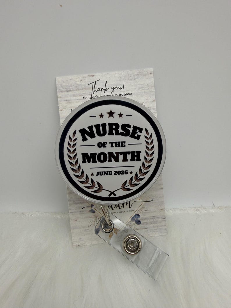 以下が含まれることがあります： 白い背景に黒い文字でNurse of the MonthとJune 2026と書かれた円形のバッジリール。黒い円形の縁取りと2つの月桂樹の葉のデザインが施されています。バッジリールには、金属製のスナップが付いた透明なプラスチック製のストラップが含まれています。