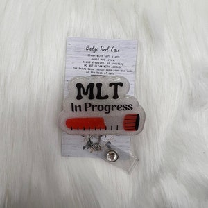 以下が含まれることがあります： 白いグリッターの背景に黒い文字で「MLT In Progress」と書かれたバッジリール。リールは、測定ツールに似た赤と黒のデザインです。ケア手順が記載されたカードがリールの後ろにあります。
