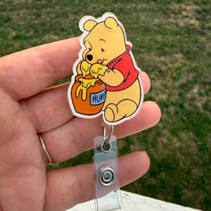 May include: A badge reel featuring a yellow bear wearing a red shirt, eating honey from an orange pot labelled HUNY. The bear is attached to a clear plastic retractable cord mechanism with a metal snap closure and a clear vinyl strap for identification.