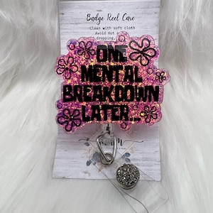 以下が含まれることがあります： ピンクのグリッターバッジリール。黒い文字で「ONE MENTAL BREAKDOWN LATER...」と書かれ、黒い輪郭のフラワーモチーフが周囲に配置されています。バッジリールは、お手入れ方法が記載された白い台紙に取り付けられており、透明なプラスチックストラップが付いています。