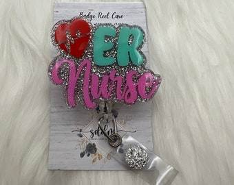 Op de afbeelding: Een badge reel met de woorden "ER Nurse" in roze, teal en zilveren glitter. Het ontwerp bevat een rood hart met een hartslaglijn. De badge reel is bevestigd aan een doorzichtige plastic houder met een decoratieve zilveren knop.