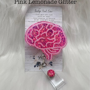 May include: A badge reel featuring a brain-shaped topper filled with pink glitter and outlined in white. The topper is attached to a clear plastic strap with a small pink glitter button. The display card includes the text Soft Pink Vinyl, Pink Lemonade Glitter, and Badge Reel Care instructions.