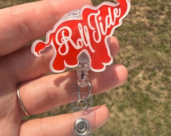 May include: A red glittery acrylic badge reel shaped like an elephant with the white cursive text Roll Tide across the center. The badge reel features a clear plastic retractable cord and a metal snap closure for attaching an identification card.