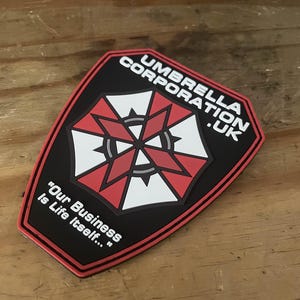 Peut inclure: Un écusson noir, rouge et blanc du logo Umbrella Corporation .UK. L'écusson présente un emblème rouge et blanc avec le texte "UMBRELLA CORPORATION .UK" et la citation "Our Business is Life Itself..."