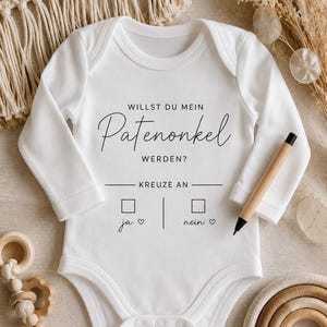May include: A white long-sleeved baby bodysuit featuring black printed text that reads WILLST DU MEIN Patenonkel WERDEN? KREUZE AN, with two checkboxes labeled ja and nein, each accompanied by a small heart symbol, and a wooden pen resting on the fabric.