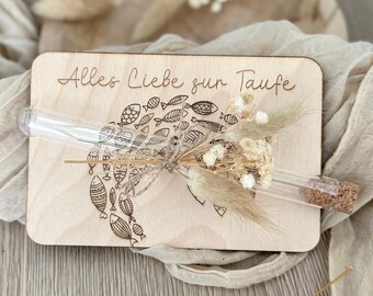 Puede incluir: Una tarjeta de madera con el texto "Alles Liebe zur Taufe" y un pequeño tubo de vidrio lleno de flores secas. El tubo está unido a la tarjeta con una cuerda.