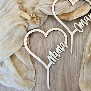 Może przedstawiać: Dwa drewniane, sercowate ozdoby do tortu z napisami "Mama" i "Oma". Ozdoby leżą na beżowym tle z tkaniny.