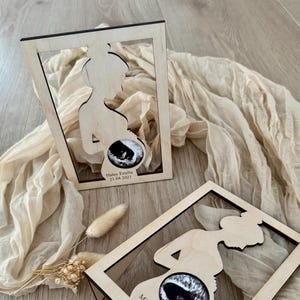 Op de afbeelding: Twee houten frames met een silhouet van een zwangere persoon die een echografiebeeld vasthoudt. Het bovenste frame heeft de tekst "Haley Emilia 21.04.2021" en het onderste frame heeft de tekst "Mila Luisa 17.02.2020".