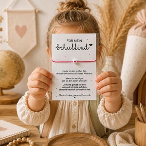 May include: A white rectangular card featuring the German text "FÜR MEIN Schulkind" and a poem, with a thin pink string bracelet attached across the center. The bracelet includes a small white bead with a tiny red heart in the middle of the string.