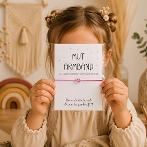 May include: A pink string bracelet with a small pink smiley face bead, presented on a white card featuring the German text MUT ARMBAND, ALLES WAS DU BRAUCHST STECKT BEREITS IN DIR, and Dein Lächeln ist deine Superkraft with a small heart symbol.
