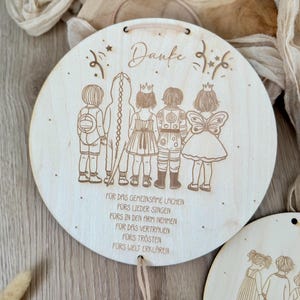 May include: A round wooden plaque with a laser-engraved design of five children in costumes. The plaque has a heart-shaped tag with the name "Anna" engraved on it. The text on the plaque reads "Danke" and "FOR DAS GEMEINSAME LACHEN FORS LIEDER SINGEN F&Uuml;RS IN DEN ARM NEHMEN FOR DAS VERTRAUEN F&Uuml;RS TR&Ouml;STEN FORS WELT ERKLAREN".