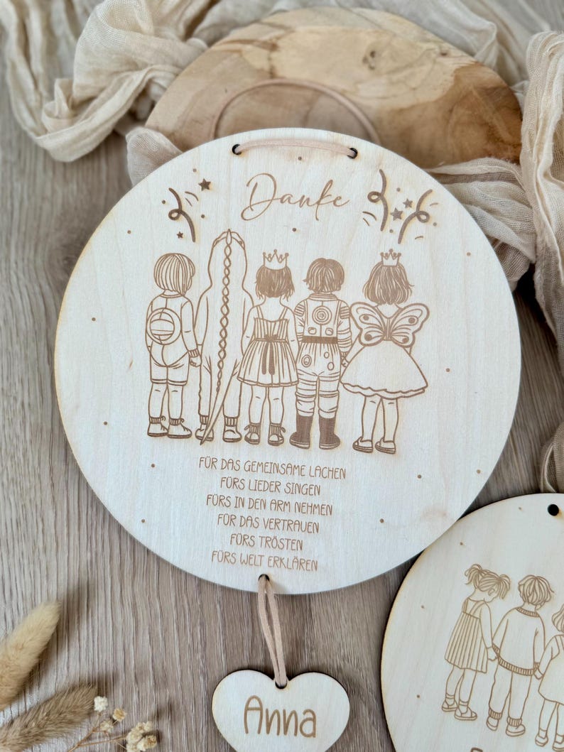 May include: A round wooden plaque with a laser-engraved design of five children in costumes. The plaque has a heart-shaped tag with the name "Anna" engraved on it. The text on the plaque reads "Danke" and "FOR DAS GEMEINSAME LACHEN FORS LIEDER SINGEN F&Uuml;RS IN DEN ARM NEHMEN FOR DAS VERTRAUEN F&Uuml;RS TR&Ouml;STEN FORS WELT ERKLAREN".