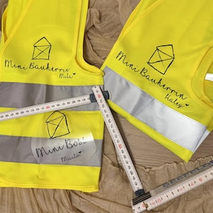 May include: Bright yellow safety vests with reflective silver stripes and the text "Mini Bauherrin" and "Mini Boss" in black script. Each vest has a small house graphic. Folding rulers are placed near the vests.