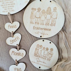May include: Wooden heart-shaped ornaments with names Anna, Steffi, Monika, and Elke hanging from a string. Three round wooden signs with illustrations of children and the text "Erzieherinnen mit Herz" and "Danke. Dass ihr uns beim wachsen geholfen habt".