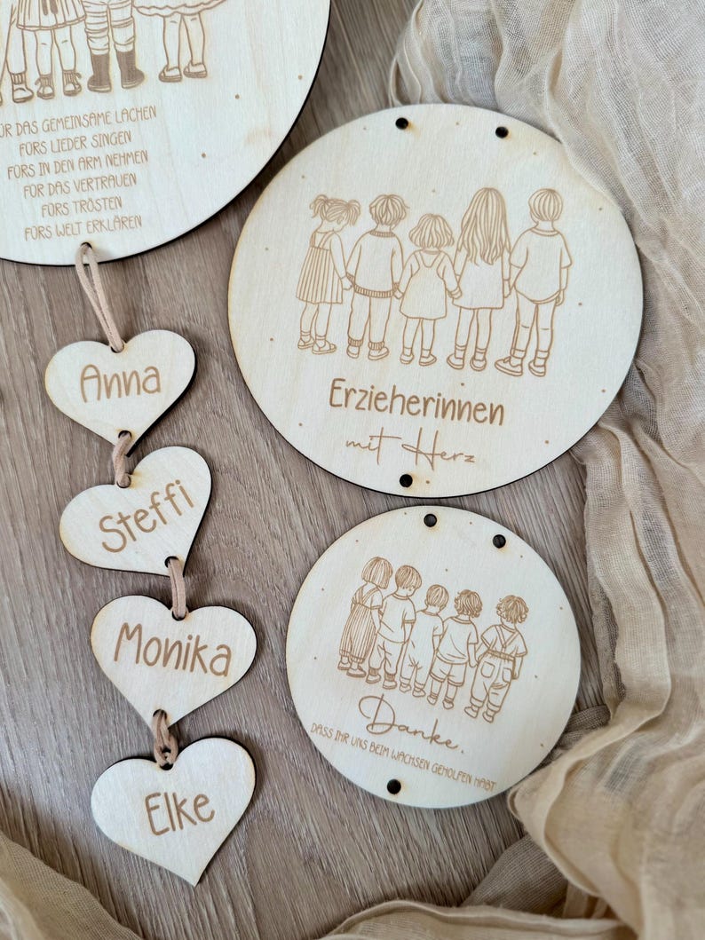 May include: Wooden heart-shaped ornaments with names Anna, Steffi, Monika, and Elke hanging from a string. Three round wooden signs with illustrations of children and the text "Erzieherinnen mit Herz" and "Danke. Dass ihr uns beim wachsen geholfen habt".
