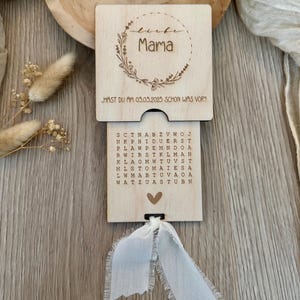 Op de afbeelding: Een houten doos met een schuifdeksel. Het deksel heeft een ontwerp met een bloemkrans met het woord "Mama" erin. De doos heeft een woordzoeker met de tekst "...HAST DU AM 05.05.2025 SCHON WAS VOR?" op de voorkant.