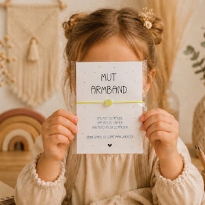 May include: A yellow string bracelet with a small smiley face bead, presented on a white card featuring the German text MUT ARMBAND, HAB MUT ZU FRAGEN, HAB MUT ZU LACHEN, HAB MUT, FEHLER ZU MACHEN, DENN GENAU SO LERNT MAN WACHSEN, decorated with small black dots.
