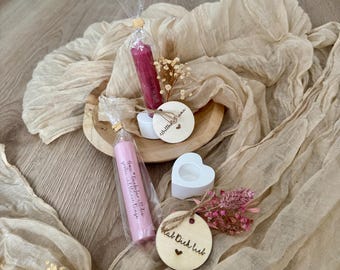 May include: Two pink candles with wooden tags that say "little dreams" and "Hab Dich Lieb". The candles are wrapped in clear cellophane and are sitting on a wooden surface with a white heart-shaped candle holder.