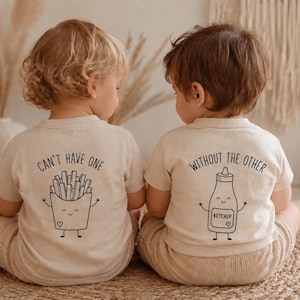 Op de afbeelding: Twee beige T-shirts met korte mouwen gedragen door kinderen, gezien vanaf de achterkant. Het linker shirt heeft een afbeelding van frietjes met de tekst "CAN'T HAVE ONE". Het rechter shirt heeft een ketchupfles met de tekst "WITHOUT THE OTHER".