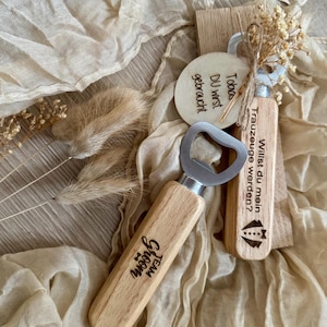 May include: Two wooden bottle openers with engraved text. One opener says "Team Groom" and the other says "Willst du mein Trauzeuge werden?" which translates to "Will you be my best man?"  A small wooden circle with the text "Du wirst gebraucht" which translates to "You are needed" is also included.