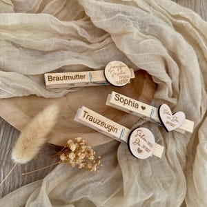 Puede incluir: Pinzas de ropa de madera con texto grabado y detalles decorativos de madera. Las pinzas presentan nombres y frases como "Brautmutter" y "Sophia". También se aprecia un adorno en forma de corazón y detalles florales.