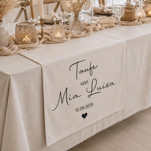 May include: A beige fabric table runner featuring black printed text that reads Taufe von Mia Luisa, the date 15.09.2026, and a small black heart icon. The runner is draped over a matching beige tablecloth, creating a clean and neutral display.