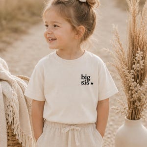 Könnte beinhalten: Ein cremefarbenes Kurzarm-T-Shirt mit dem schwarzen Schriftzug "big sis" und einem kleinen schwarzen Herzsymbol auf der linken Brustseite. Das Shirt hat einen Rundhalsausschnitt und ein schlichtes, klares Design für den Alltag.