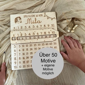 Könnte beinhalten: Holz-Multiplikations-Tabelle mit Schieberegler zum Lernen von Mathematik. Die Tabelle zeigt die Zahlen 1 bis 10 und den Namen "Mila" oben. Der Text "Über 50 Motive + eigene Motive möglich" ist ebenfalls auf der Tabelle zu sehen.
