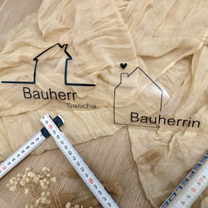 Könnte beinhalten: Zwei durchsichtige Acryl-Schilder in Hausform mit schwarzen Umrissen und Text. Das Schild links lautet "Bauherr Sascha" und das Schild rechts lautet "Bauherrin".