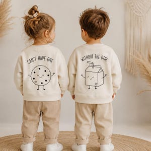 Könnte beinhalten: Zwei cremefarbene Langarm-Sweatshirts mit schwarzen Strichgrafiken. Das linke Sweatshirt zeigt einen lächelnden Keks mit dem Text CAN'T HAVE ONE, das rechte einen Milchkarton mit dem Text WITHOUT THE OTHE auf der Rückseite.