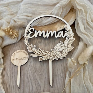 Può includere: Topper per torta in legno con una ghirlanda di fiori e il nome "Emma" in un font corsivo. Un topper circolare in legno separato dice "Zur Kommunion am 16.03.2025".