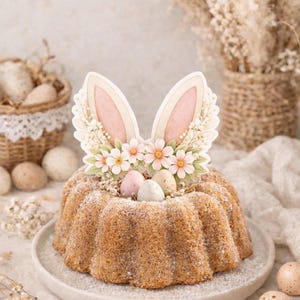 Op de afbeelding: Een tulbandcake versierd met een konijnenoortopper, bloemen en pastelkleurige eieren. De cake is bestrooid met poedersuiker en staat op een licht bord, perfect voor Pasen. De cake heeft een diameter van ongeveer 20 cm.