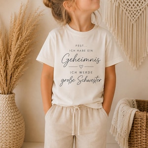 Könnte beinhalten: Ein cremefarbenes Kurzarm-T-Shirt mit schwarzem Aufdruck PSST... ICH HABE EIN Geheimnis, gefolgt von einem kleinen Herzsymbol und dem Text ICH WERDE große Schwester. Das T-Shirt wird mit einer hellbeigen Hose mit Kordelzug kombiniert.