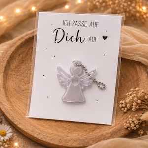 May include: A clear, translucent angel-shaped charm with a silver-toned ball chain attached to a white rectangular card. The card features the black printed text "ICH PASSE AUF Dich AUF" with a small black heart symbol and scattered black dots.