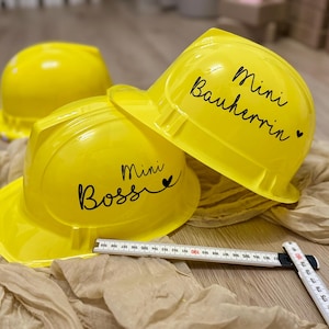 May include: Bright yellow construction helmets with black script that reads "Mini Boss" and "Mini Bauherrin". A folding ruler is partially visible. The helmets are on a beige fabric.