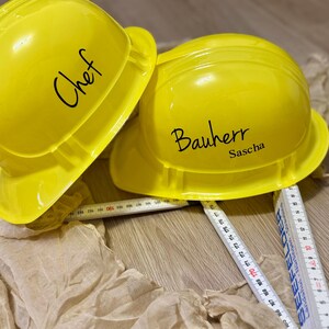 May include: Two bright yellow construction helmets, one labeled "Chef" and the other "Bauherr Sascha", rest on a wooden surface. A folding ruler is partially visible. The scene suggests a construction or culinary theme.