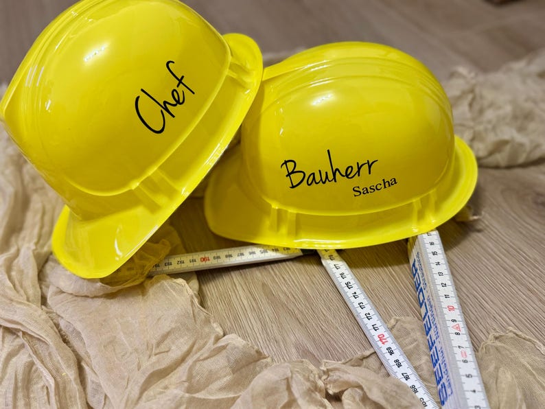 May include: Two bright yellow construction helmets, one labeled "Chef" and the other "Bauherr Sascha", rest on a wooden surface. A folding ruler is partially visible. The scene suggests a construction or culinary theme.