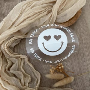 Può includere: Un adesivo circolare trasparente con una faccina sorridente bianca con occhi a forma di cuore e una bocca curva. Il testo "SEI FRECH - WILD UND WUNDERBAR" è stampato attorno alla faccina sorridente. L'adesivo è su una superficie di legno con tessuto beige.