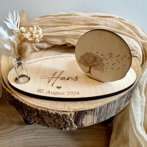Peut inclure: Une plaque en bois gravée du nom "Hans" et de la date "02. Août 2024". Un petit flacon en verre contenant des fleurs séchées est placé dans une découpe sur la plaque. Un cercle en bois gravé d'un arbre et d'oiseaux est fixé à la plaque.