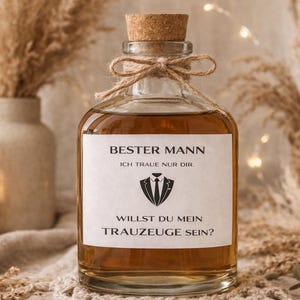 Puede incluir: Una botella de vidrio transparente llena de líquido ámbar, tapada con un corcho y atada con hilo. Una etiqueta blanca muestra texto y un gráfico de esmoquin. La botella forma parte de un tema de boda.