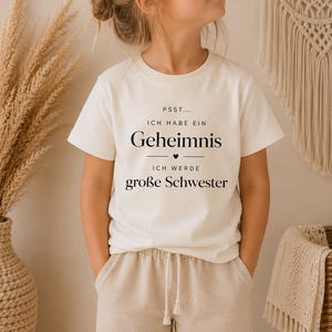 Könnte beinhalten: Ein cremefarbenes Kurzarm-T-Shirt mit schwarzem Aufdruck: PSST... ICH HABE EIN Geheimnis ICH WERDE große Schwester, inklusive eines kleinen Herzsymbols, das mittig zwischen den Textzeilen auf der Vorderseite des Kleidungsstücks platziert ist.