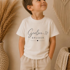 Può includere: Una t-shirt a maniche corte color crema con la scritta nera "Großer BRUDER", una piccola icona a forma di cuore sopra la parola "Großer" e un'icona a forma di cuore centrata tra due linee orizzontali sotto la parola "BRUDER".