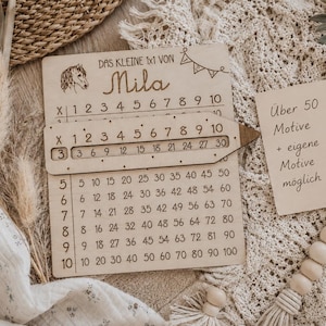 May include: A rectangular wooden multiplication board featuring the text 'DAS KLEINE 1x1 VON Mila' and a horse illustration. It includes a sliding pencil-shaped wooden marker and a separate rectangular card with the text 'Über 50 Motive + eigene Motive möglich' and a heart symbol.