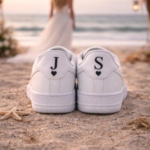 Puede incluir: Zapatillas blancas con las iniciales "J" y "S" y un símbolo de corazón en la parte trasera, sobre una playa de arena. Una novia con un vestido blanco está al fondo, cerca del mar. Calzado de boda.