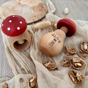 Könnte beinhalten: Holzspielzeug in Pilzform mit roten Oberteilen und weißen Punkten, zusammen mit einem Holzbrett und Walnüssen. Ein Pilz trägt den Namen "Haley" und eine Hasenillustration. Das Spielzeug liegt auf einem beigen Tuch.
