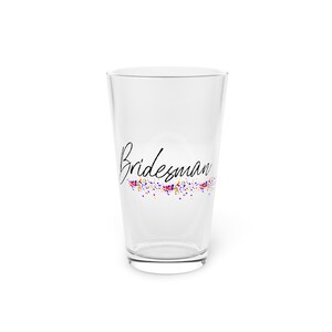 Op de afbeelding: Een helder pintglas met het woord "Bidesman" in zwart schrift. Onder het woord staat een kleurrijk confetti-ontwerp in rood, blauw, geel en paars. Het glas is leeg en staat op een witte ondergrond.