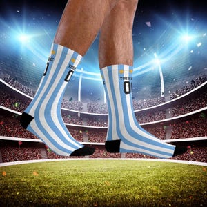 Puede incluir: Un par de calcetines a rayas azules y blancas con un número "0" negro y dorado y el texto "YOUR NAME" en ellos. Los calcetines los lleva una persona de pie en un estadio de fútbol.