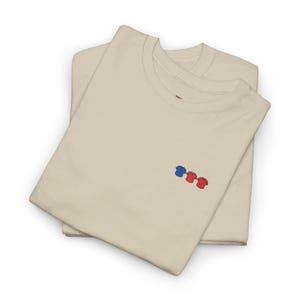 Puede incluir: Dos camisetas crema dobladas con un pequeño diseño bordado de tres camisas en miniatura en azul, rojo y rojo. Las camisetas están hechas de un material suave y están cuidadosamente apiladas.