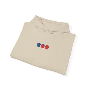 Puede incluir: Sudadera con capucha beige con cordón. La parte delantera de la sudadera presenta un diseño bordado de tres camisetas pequeñas en azul, rojo y rojo. La sudadera está doblada.
