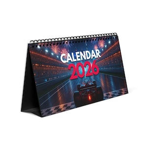Puede incluir: Un calendario de escritorio con encuadernación en espiral negra. La portada presenta un coche de carreras en una pista con las palabras "CALENDAR 2026" en blanco y rojo. El fondo muestra un estadio con fuegos artificiales.